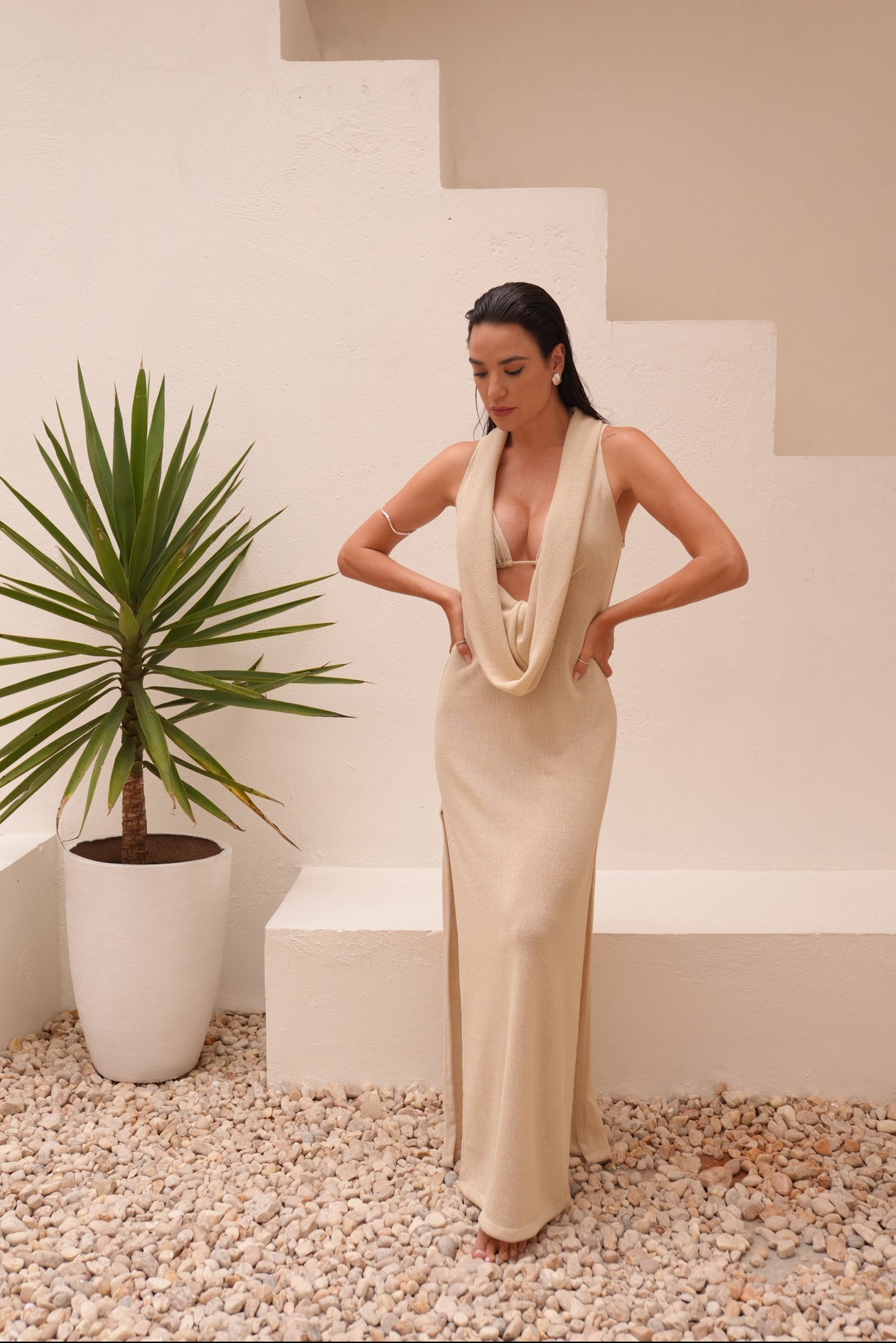 Woman wearing off-white knit maxi dress with draped neckline and open back, standing by earthy-toned archway.