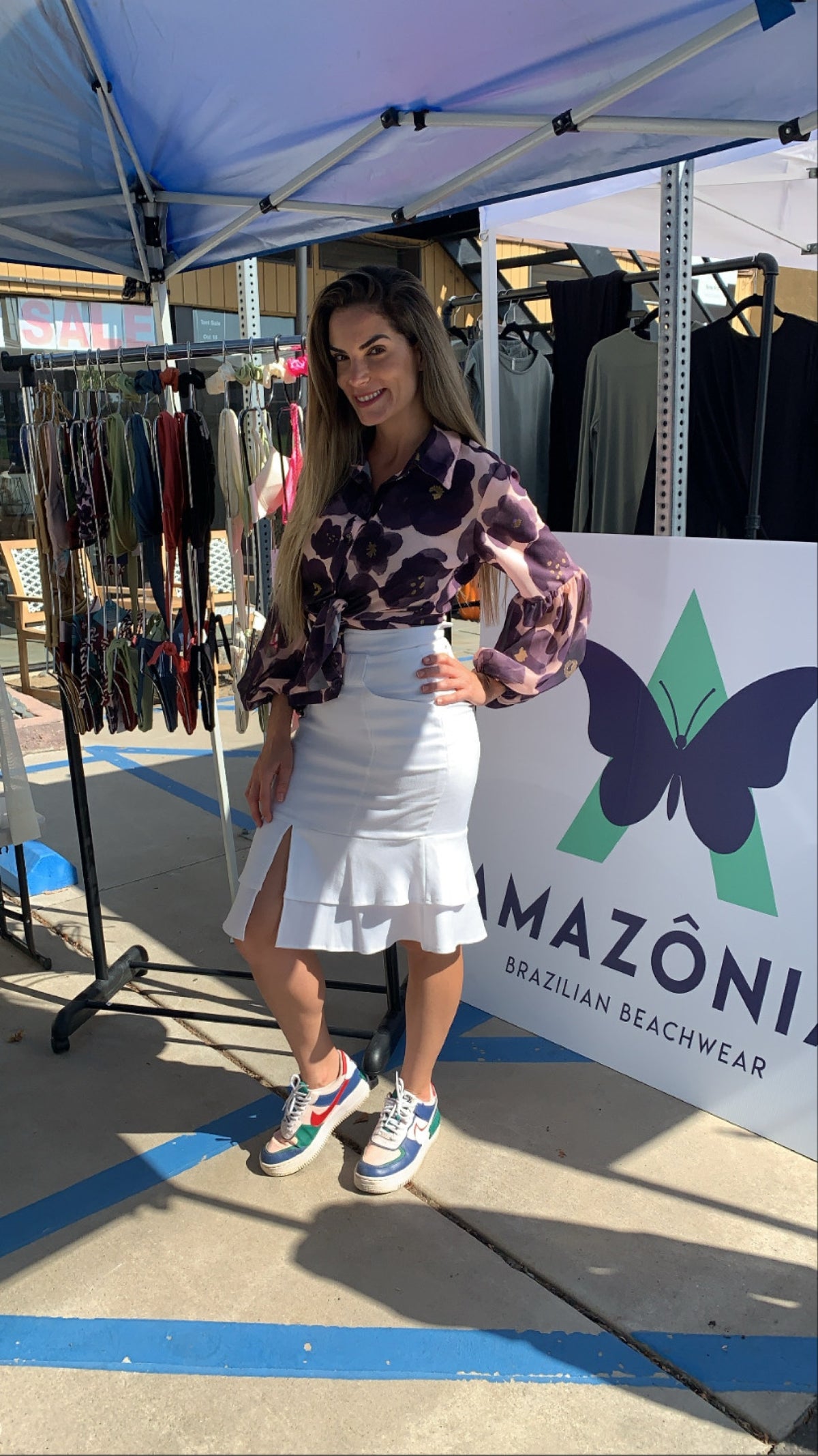 Woman at an outdoor pop-up tent next to an AMAZÔNIA Brazilian Beachwear butterfly banner with clothing racks