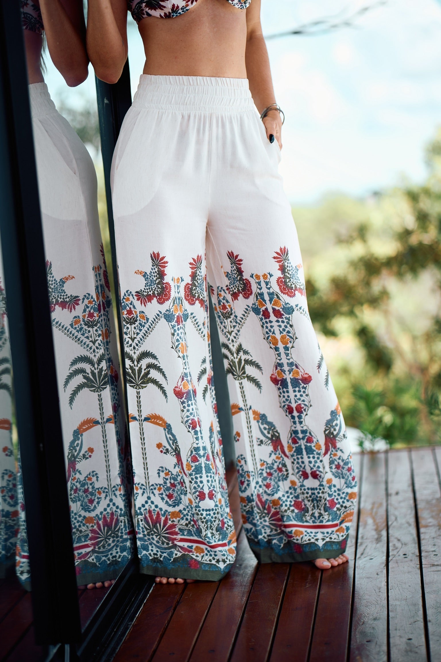 Woman standing barefoot wearing tropical white pants with colorful Amazon-inspired print, reflected in glass.
