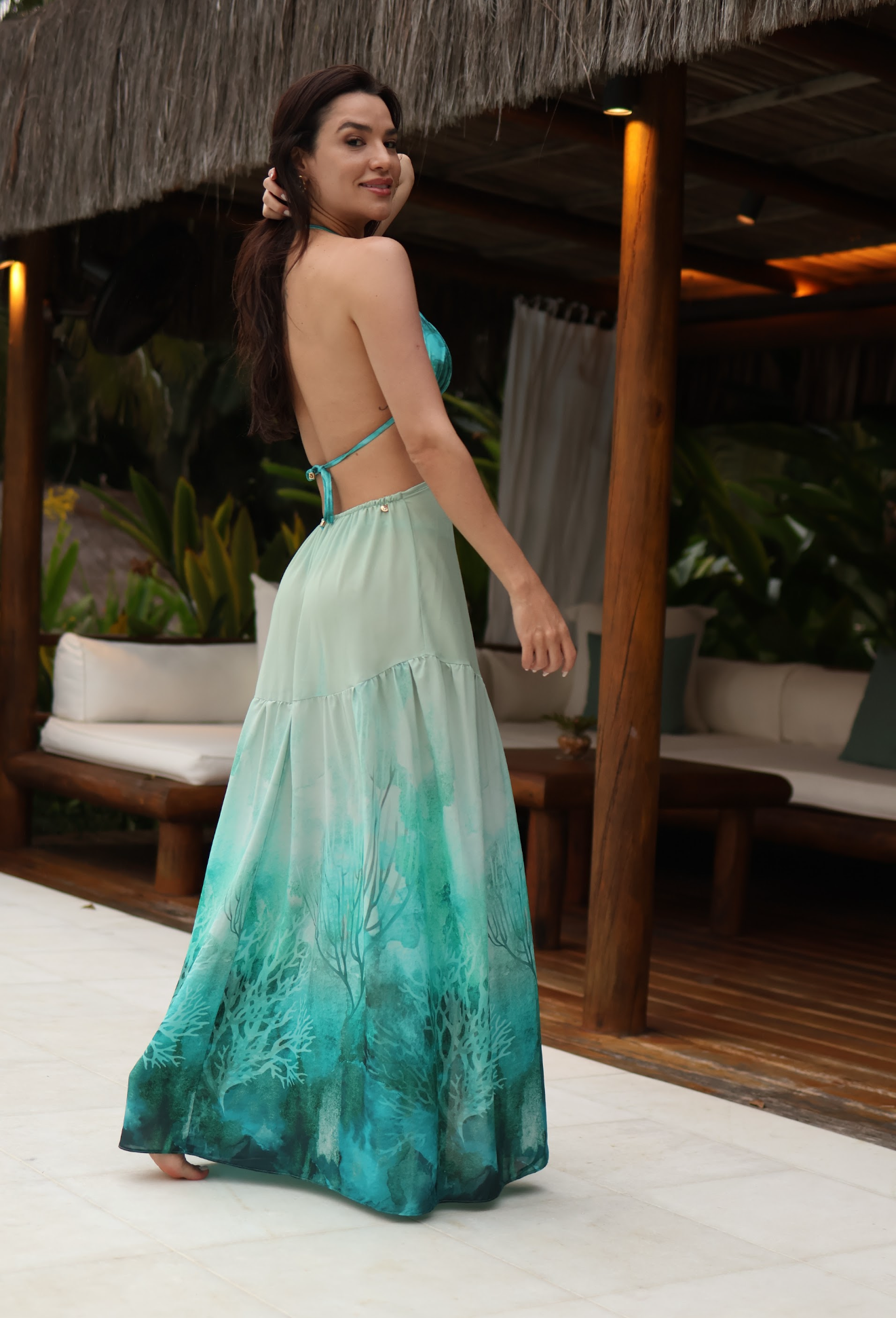 A woman wearing a flowing maxi dress with a gradient design transitioning from light mint green to teal and turquoise, standing in an outdoor patio setting.