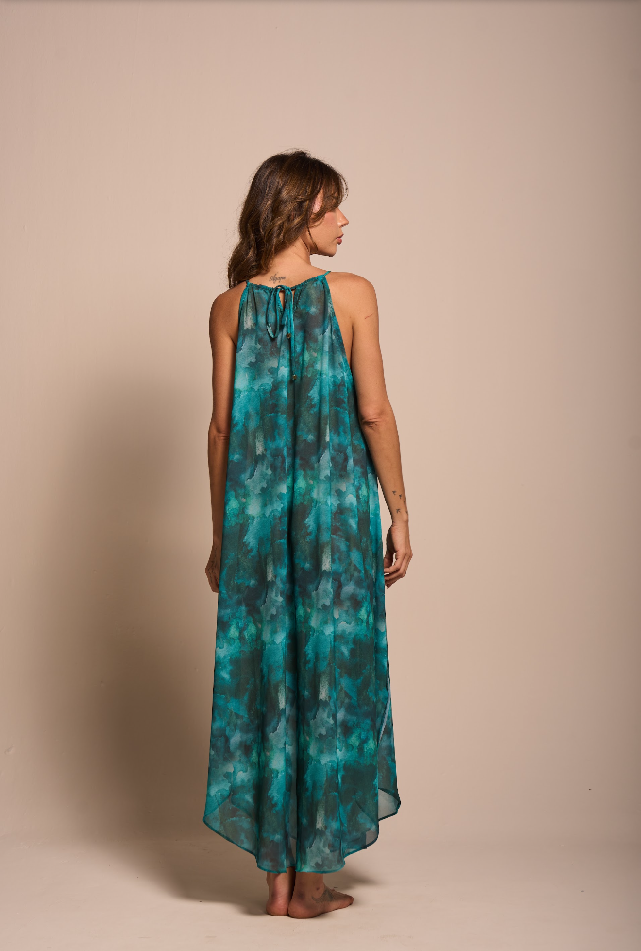 A woman stands with her back to the camera wearing a sleeveless, high-neck, floor-length maxi dress with a teal and green watercolor tie-dye pattern, set against a solid beige studio background.