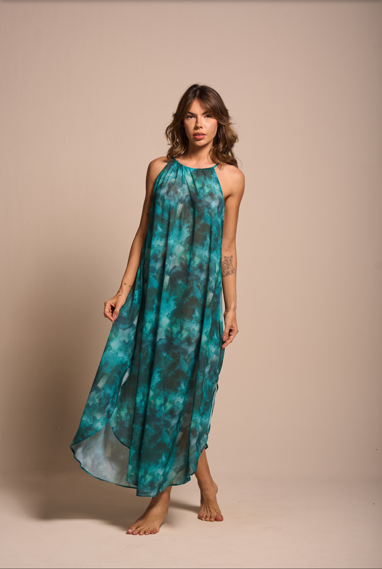 A woman modeling a sleeveless halter-neck maxi dress with a teal and black tie-dye pattern, standing against a solid beige studio backdrop.