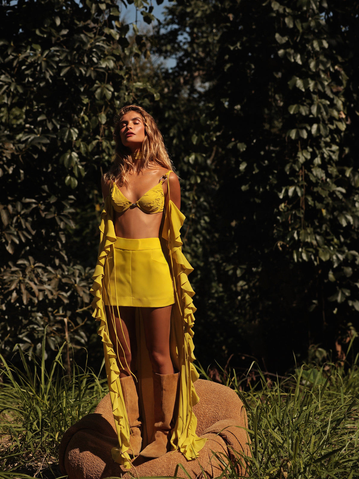 A woman stands outdoors in a grassy, wooded setting wearing a bright yellow two-piece outfit with a ruffled long-sleeve cover-up and tan knee-high boots.