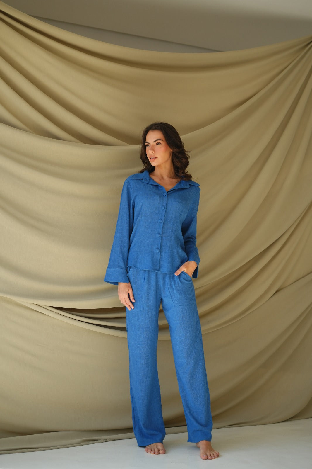 Model wearing blue cotton resort pants from Amazônia B2R, barefoot in a soft beige draped backdrop – easy, elegant, and travel-ready