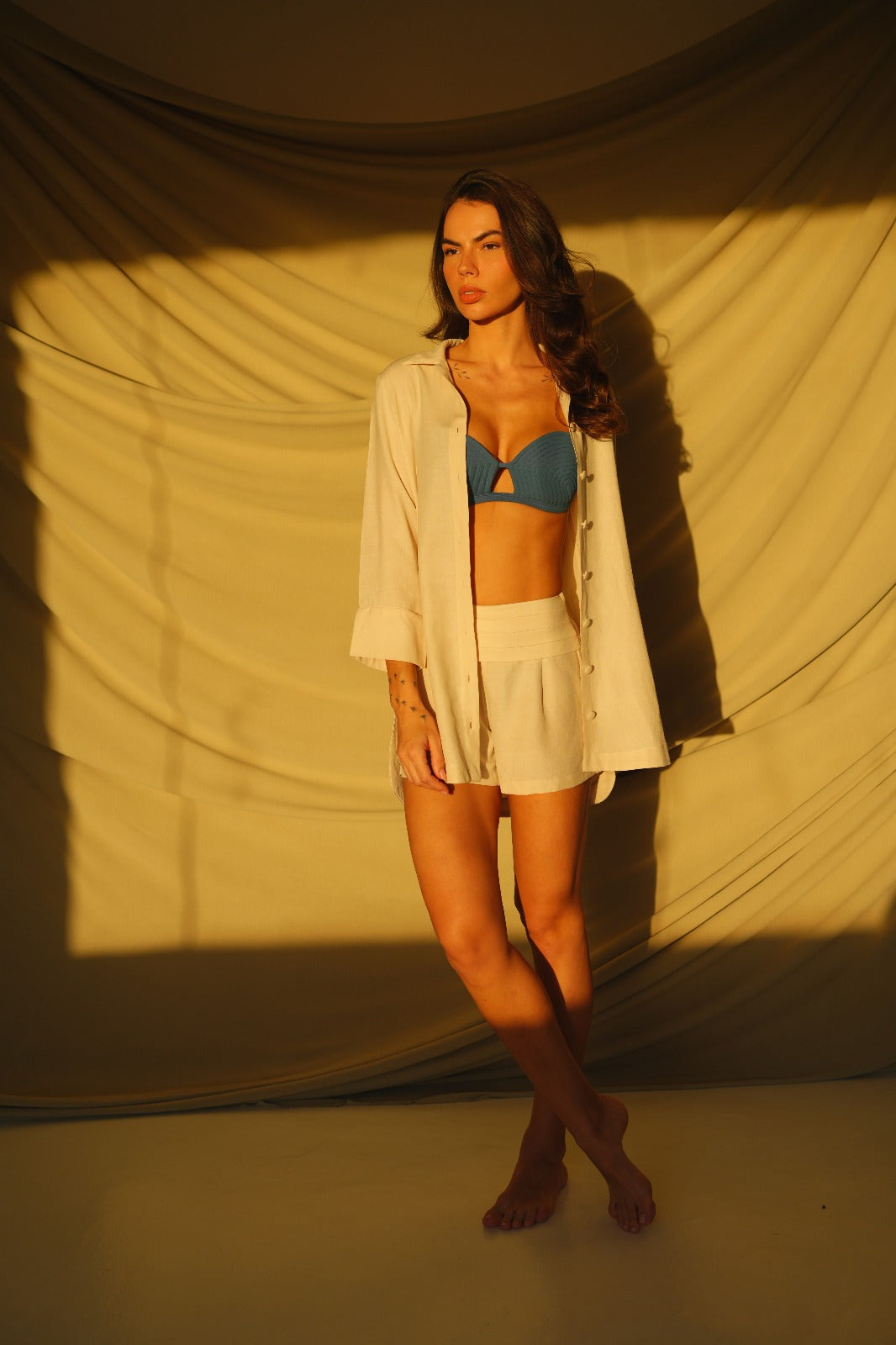 Model wearing sand-colored linen shorts with pleated high waist and golden button detail. Perfect for warm destinations, over bikinis or resort tops.