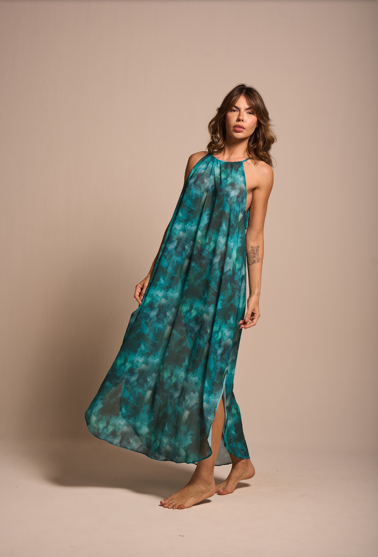 A woman modeling a sleeveless halter-neck maxi dress with a teal and turquoise watercolor tie-dye pattern, standing against a solid beige studio background.